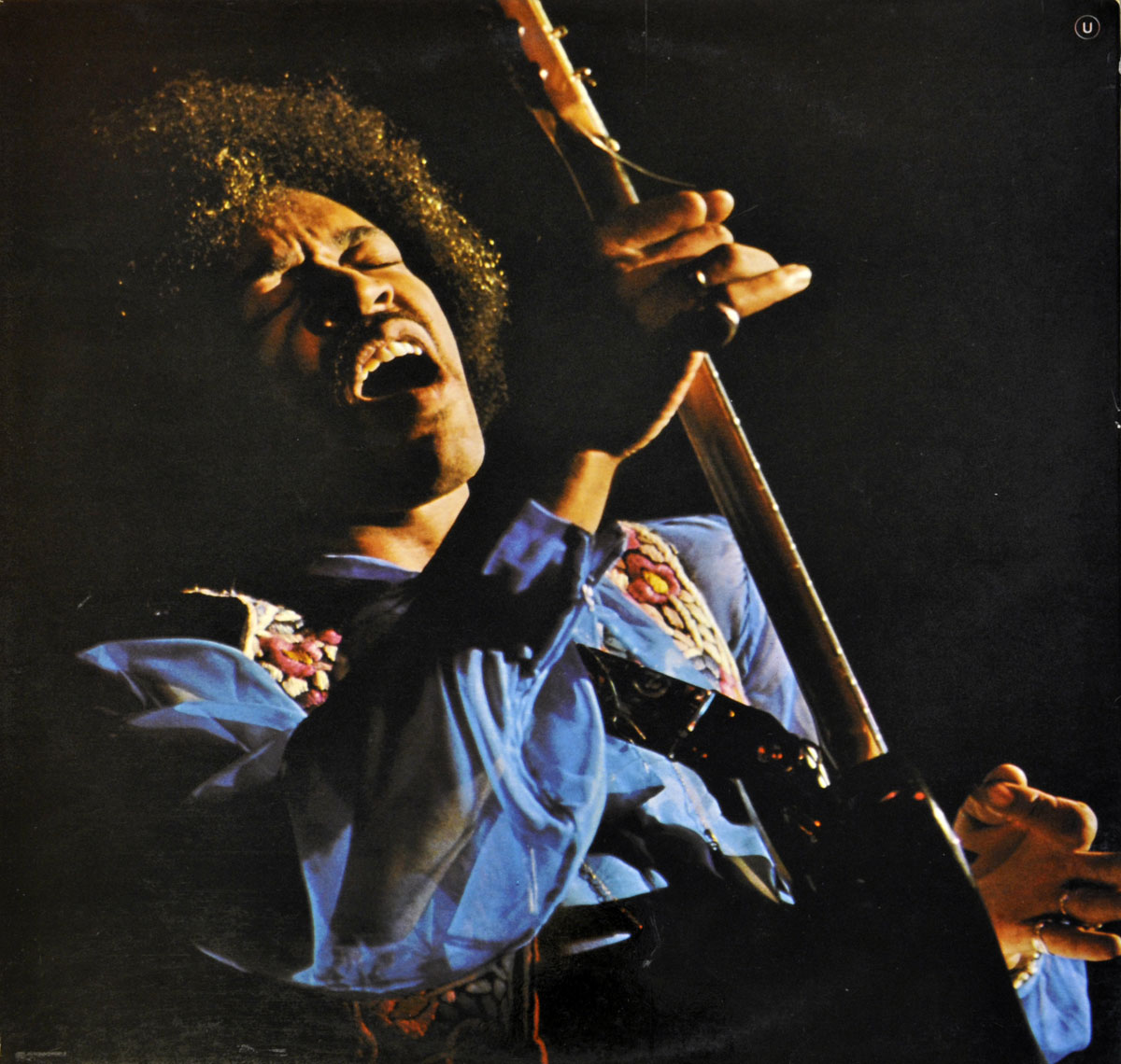 Jimi Hendrix in a powerful live performance, wearing a vibrant blue, flowing shirt adorned with intricate floral embroidery. His face is filled with intense emotion as he sings with his mouth open and eyes closed, deeply immersed in the music. He grips his guitar at a dramatic angle, fingers pressing the strings, embodying the raw energy of rock and blues. The stage lighting casts warm highlights on his afro and outstretched hand, enhancing the sense of movement and passion.